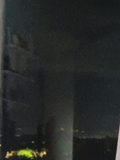This media file, captured in Kosh, Armenia, displays a highly dark and indistinct scene, primarily in shades of black and dark gray. The setting appears to be a dimly lit or unlit environment, possibly indoors or an outdoor space at night, given the pervasive low light.
On the right side of the frame, a vertical, dark object or structure is visible. This object features notable patches of bright, yellowish-green material, which could be moss, algae, or some form of plant life growing on its surface. These green patches are the most prominent and colorful detail in the image.
In the lower central and left portions, faint and blurry dark shapes are present against the equally dark background. These shapes are too ambiguous to identify definitively but may suggest furniture, architectural features, or other obscured objects. A horizontal line near the bottom edge could indicate a floor level or the base of a wall.
No people or discernible specific activities are visible. The low resolution and extreme darkness prevent any clear identification of objects, details, or emotions. There is no visible text. The overall impression is one of obscurity and poor visibility, with a subtle texture throughout, possibly from dust or surface imperfections.