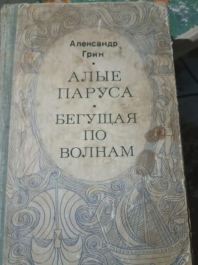 This is a close-up, top-down view of the cover of a book titled "Алые Паруса" (Scarlet Sails) by Александр Грин (Alexander Grin). The cover features an ornate blue and grey line drawing against a faded, textured background. The design depicts a nautical theme with sailing ships, waves, and stylized figures of a woman and a man. The woman, on the left, is depicted with her arms raised, seemingly in a celebratory or expectant pose, while the man on the right is also looking upwards with his arm raised. The central text is arranged in several lines, with the author's name at the top, followed by the title in larger font, and then a subtitle. The overall aesthetic suggests a vintage or classic storybook. The text is in Russian. The book cover appears worn, with some stains visible, particularly on the right side near the drawing of the man. The book is bound in a light blue cloth. The scene depicted is a symbolic representation of the story, likely evoking themes of dreams, hope, and adventure at sea.