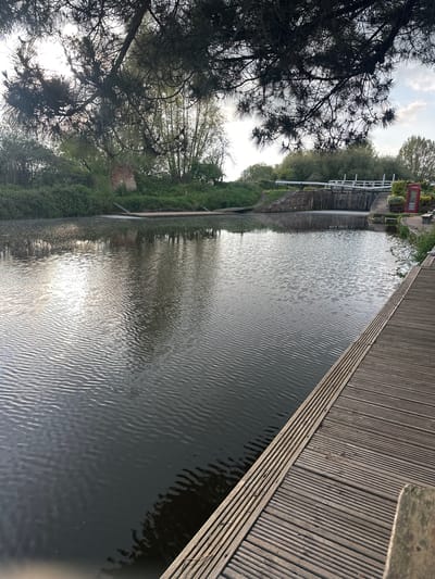 The image depicts a canal in Exeter, United Kingdom. A wooden dock runs along the right side of the frame, constructed of parallel planks. Water fills the majority of the frame, exhibiting a rippled surface reflecting the sky. Along the left bank are trees and vegetation. A red telephone box is visible further up the canal, near a lock system with white gates. The sky is overcast with patches of light.