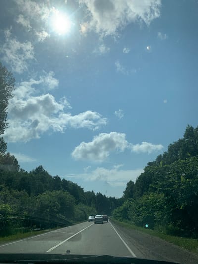 The image depicts a view from inside a vehicle traveling on a highway. The road is asphalt, with white lane markings and a guardrail visible on the left side. Dense green trees line both sides of the road, forming a canopy that extends into the distance. The sky is blue with scattered white clouds, and the sun is prominently visible in the upper left quadrant, casting a bright light. Several cars are visible on the road ahead, traveling in the same direction. The scene appears to be a rural or semi-rural setting in Russia, potentially along the Doma 78 Km highway, given the provided context.