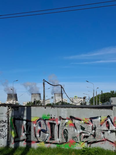 The image depicts a section of a large, grey concrete wall covered in graffiti. The graffiti on the main section of the wall features large, stylized lettering in shades of grey, black, red, and yellow. To the left, there is additional graffiti in black and white. The wall is situated in front of a grassy area and appears to be part of an industrial or urban landscape. In the background, three large cooling towers from a power plant are visible, emitting plumes of steam into a clear blue sky. Several apartment buildings and streetlights are also present in the background, suggesting an urban setting within Novoivanovskoe, Russia. Power lines are strung across the top of the frame.