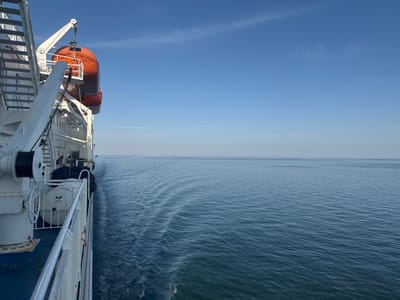 The image shows the side of a large ferry or ship sailing on a calm, blue sea under a clear sky with a few wispy clouds. The sun is shining, suggesting it is daytime. The perspective is from the deck of the vessel, looking forward and to the right, with the wake of the ship visible in the water.

Prominently displayed on the upper deck is an orange lifeboat, suspended by a davit. Text on the lifeboat reads "ROBIN HOOD EMDEN". Below the lifeboat, the deck is equipped with railings, a large winch with a coiled rope, and other equipment. A few people are visible on the deck, appearing to be passengers or crew, sitting and observing the surroundings.

The sea is a deep blue, with gentle ripples indicating smooth sailing. In the far distance, a faint, hazy line suggests land on the horizon, possibly indicating the vessel is approaching or leaving a port. The overall mood is peaceful and serene, capturing a moment of travel by sea. The image conveys a sense of journey and the vastness of the ocean.