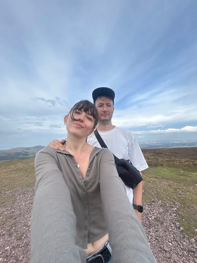 Two individuals, a woman in the foreground and a man behind her, are taking a selfie on a rocky, grassy hillside with a cloudy sky. The woman has dark hair with bangs and is wearing a light brown, button-up cardigan and jeans. She is smiling and looking at the camera, with her arm around the man's shoulder. The man is wearing a white t-shirt, a black baseball cap, and glasses. He has a black bag slung across his body. The background shows rolling hills and a distant view of what appears to be a town or city. The location is identified as being in the vicinity of Penicuik, United Kingdom.