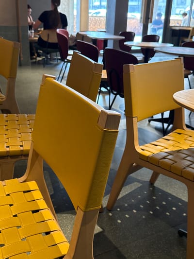 The image shows a series of chairs in what appears to be a cafe or restaurant setting, likely located in Hongkou District, China, based on possible contextual clues (though this cannot be definitively confirmed from the image alone). The chairs are made of light-colored wood with yellow leather or vinyl seats and woven backrests. The camera angle is close-up, focusing on the details of the chairs. In the background, through a window, we can see a blurred cityscape. A person is visible, working behind a counter. The light suggests it could be daytime. Other tables and chairs, some with red seats, are partially visible, indicating a seating area. The flooring appears to be dark and patterned.