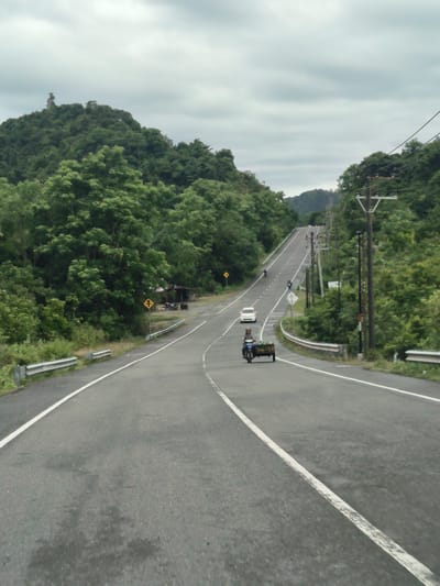 An overcast day frames a two-lane asphalt road winding through a lush, green landscape. Hills covered in dense vegetation rise on either side of the road, with some appearing steeper and more rugged than others. Power lines run along the right side of the road, supported by wooden poles.

In the foreground, the road curves gently to the right. A person is riding a motorized tricycle loaded with green produce in the right lane, heading away from the camera. Further up the road, a white car is visible in the left lane, also moving away. Two motorcycles are further up the incline, one in each lane. The road stretches into the distance, disappearing over a crest.

On the left side of the road, a metal guardrail runs alongside the shoulder. Yellow traffic signs are posted at intervals along the roadside. To the left of the road, near a slight bend, a small, open-air shelter with a dark roof is visible, with two motorcycles parked nearby.

The overall scene depicts a rural or semi-rural road in Pulot, Indonesia, characterized by its verdant surroundings and moderate traffic.