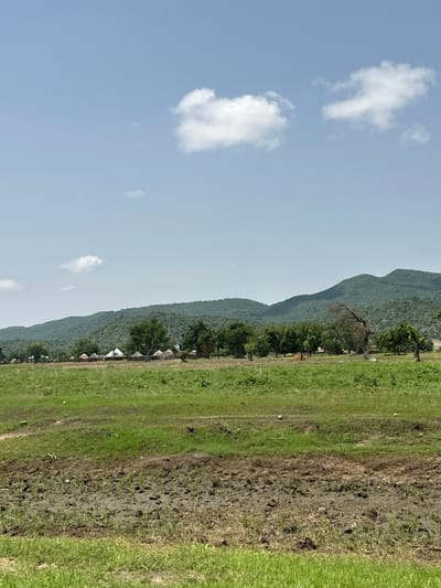 A wide shot captures a rural landscape under a clear blue sky with scattered white clouds. In the foreground, a muddy patch of earth interrupts a vibrant green grassy field. Further back, a cluster of thatched-roof huts forms a small village, nestled amongst scattered trees and foliage. Beyond the village, a lush, verdant mountain range dominates the background, its slopes covered in dense trees. The scene appears serene and undeveloped, suggesting a remote, agricultural setting. This location is in the city of Atawa, Nigeria.
