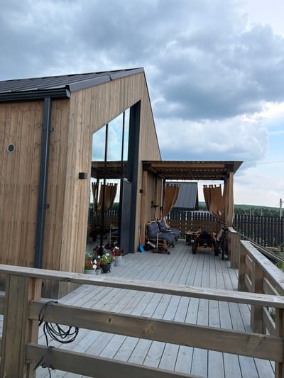 The image shows a modern wooden house with large windows and a wooden deck. On the deck, there is a covered seating area with two chairs, a small table, and some curtains. There are also two vehicles, a go-kart and a bicycle, and some potted plants. A fence is visible in the background, along with a landscape of trees and a cloudy sky. The scene is set in Maloe Sedelnikovo, Russia.