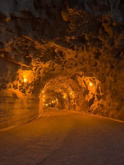 The image depicts a tunnel illuminated by a series of evenly spaced small lights. The tunnel's left wall is rough and uneven, consisting of what appears to be exposed rock or earth. The right wall is smooth and seemingly constructed from concrete or similar material. The tunnel floor is paved and appears to be slightly damp. The location is Ponta do Sol, Portugal. No people are visible in the image. The scene is quiet and still, emphasizing the structure of the tunnel itself.