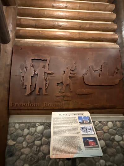 The image shows a historical exhibit about the Underground Railroad, located in New Harmony, Indiana, United States. The exhibit features a large, rust-colored metal relief depicting a wagon and figures, with the words "Freedom Bound" inscribed below. To the right of the relief is an informational sign with text and three small photographs. The sign discusses the Underground Railroad, its routes, and its significance. The background of the exhibit is constructed from stacked logs and a stone wall. The overall presentation suggests a museum or historical site display.