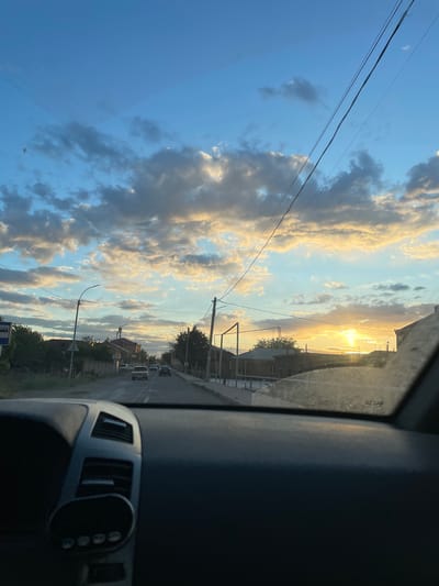 The image, taken from inside a vehicle, shows a street in Aratashen, Armenia at sunset. The sky is filled with clouds illuminated by the setting sun. Power lines are visible against the sky. Along the street, buildings, trees, street lights, and parked vehicles can be seen. The dashboard of the vehicle is partially visible in the foreground.