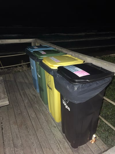 A row of recycling bins lines a wooden deck. The bins, in hues of blue, green, yellow, and black, are positioned against a railing of sorts. A black trash bag is draped over the top bin. The "Vagos" logo is visible on one of the bins. To the left, darkness dominates, suggesting a nighttime setting. The wooden planks of the deck lead the eye towards the right side of the image, where a small patch of greenery is visible.