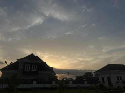 The image depicts a residential area in Pakuro, Nigeria, during a cloudy sunset. Two houses are visible: a larger, darker house on the left with a distinctive multi-level roof and several windows, and a smaller, lighter colored house on the right with a simpler roofline and windows. Both structures appear to be modern residences. In the foreground, manicured green grass and small palm trees are present. A white fence with black decorative elements separates the properties from the grassy area. Utility poles and power lines are visible in the background, suggesting an established neighborhood. The sky is filled with dramatic, layered clouds, with a warm orange glow emanating from the horizon where the sun is setting. The overall atmosphere is calm and serene, characteristic of the end of the day. No people are visible in the image.