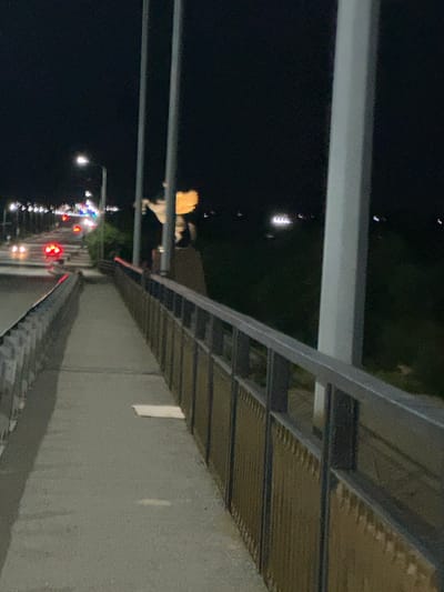 This is a nighttime, slightly blurry, horizontal shot from a pedestrian walkway on a bridge. The walkway is made of concrete and has a metal railing with a gold-colored mesh filling along the right side. A large, white vertical pillar is prominent on the right side of the frame. In the distance, cars with illuminated headlights and taillights are visible on a road running parallel to the walkway. There appear to be streetlights and other distant lights indicating a populated area. A statue or monument, partially lit with a golden glow, is visible in the background to the left of the white pillar. The scene is dark with artificial lights providing illumination.