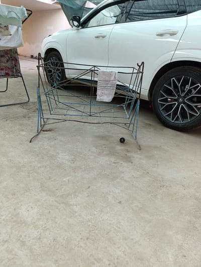 A weathered blue metal baby cradle with a folded towel rests on a concrete surface. A white car is parked behind the cradle. The scene is outdoors, possibly in a garage or open-air parking area, with a hint of an urban environment suggested by the presence of the car.