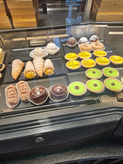 The image displays a selection of pastries in a display case, likely in a bakery or patisserie in St. Moritz, Switzerland. The pastries are arranged on black trays. In the top section, there are round tarts with yellow filling, some with "Guitron" written in black script on top, and others with bright green filling topped with a single chocolate piece. Below these are several cream-filled pastries shaped like cones, dusted with powdered sugar. There are also chocolate-covered pastries with white swirl decorations and dark chocolate pastries dusted with cocoa powder.
To the left of the pastries, stacked golden boxes with embossed designs are visible, suggesting packaged goods for sale. The overall setting appears to be indoors, with the display case reflecting the surroundings. The lighting suggests it could be daytime. There are no people visible in the frame. The context of St. Moritz, Switzerland, implies a potential setting in a resort town, possibly catering to tourists. The focus is solely on the visually appealing arrangement of the baked goods.