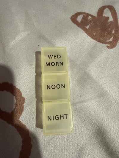 The image shows a small, translucent pill organizer with three compartments stacked vertically. Each compartment has text printed on it. The top compartment reads "WED MORN". The middle compartment reads "NOON". The bottom compartment reads "NIGHT". The pill organizer is placed on a light-colored fabric with a textured pattern, possibly a bedsheet or tablecloth, which features a faint reddish-brown drawing of a heart on the right side. The lighting in the image suggests it was taken indoors, possibly at night or in a dimly lit room, indicated by the shadow cast by the pill organizer. There are no people or any other discernible objects in the image. There is no information within the image to determine the location in the city of Manila, Philippines.