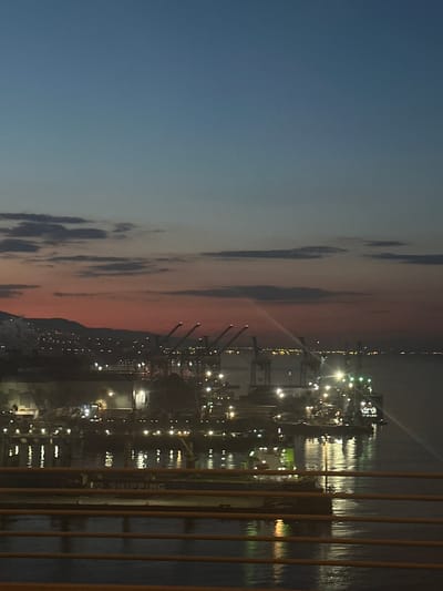 The image shows a night view of Dilovası, Turkey, likely taken from an elevated position. The sky is split, with a dark blue on the left transitioning to an orange-red hue near the horizon where the city is located. Clouds are visible in the sky. In the city, many lights illuminate what appears to be an industrial port area. Cranes stand prominently. A body of water reflects the city lights. A vertical grid-like structure, probably a railing or fence, is in the foreground.