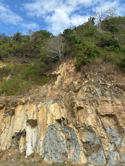 This image depicts a steep, rocky mountainside covered in lush green vegetation and scattered trees under a bright blue sky with scattered white clouds. The mountainside is characterized by exposed rock formations, varying in color from tan and brown to grey and blueish hues, suggesting different mineral compositions. The vegetation consists of dense shrubs and trees, with some bare, lighter-colored branches of trees visible, particularly towards the top of the incline. The terrain appears rugged and natural, indicative of a hillside or a quarry. The location is identified as Taliwang, Indonesia. There are no people or visible human activity in the image. The overall scene is one of natural landscape.