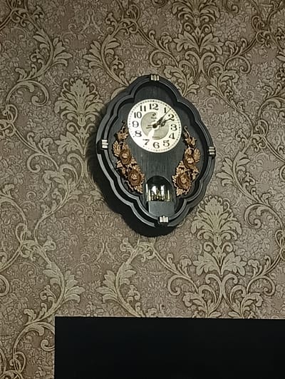 The image shows a wall clock hanging on a patterned wallpaper. The clock has a black, ornate frame and a white face with black numbers and hands. The hands indicate the time is approximately 11:32. The wallpaper has a repeating damask pattern in shades of brown and beige. There is a dark, rectangular object partially visible at the bottom of the image. The scene does not provide any specific information about the location in the city of Тайжина, Russia.