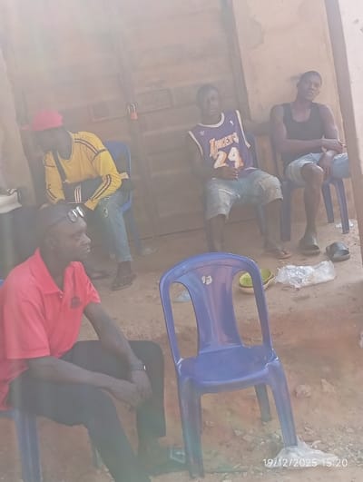 Four men are seated in chairs outdoors, likely in a rural setting. One wears a red shirt and black pants, sunglasses perched on his head, looking to the side. Another sports a yellow long-sleeved shirt and a red cap. A third man wears a Lakers jersey. The fourth man, seated furthest back, is in a black tank top. A blue plastic chair is in the foreground. The area is sandy, and there are some dishes on the ground nearby. There is a door in the background with a lock.