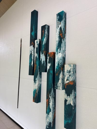 In an indoor setting with a textured white wall, a collection of abstract, rectangular art pieces are mounted. These pieces are painted with a dark teal as the base color, overlaid with textured strokes of white and hints of orange. They are arranged vertically and staggered at different heights, creating a dynamic visual effect. To the left of the art installation, a long, slender dark object with a pointed tip, resembling a spear or a staff, is mounted vertically on the wall. The floor beneath is tiled and appears to be a light neutral color. The ceiling, visible in the upper left corner, features parallel wooden slats. The overall impression is of modern interior decor. The location is identified as Unknown, Maldives.