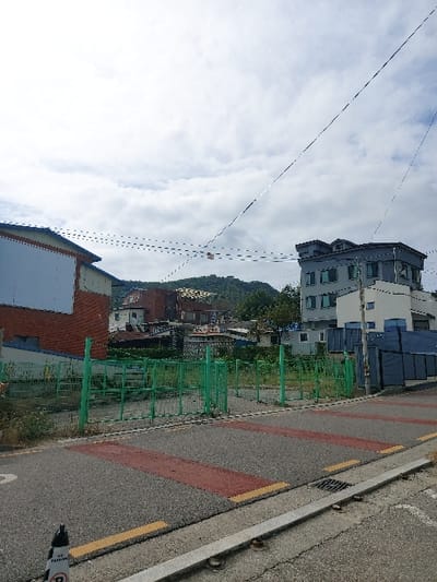 The image displays an urban-suburban street scene in 강화군, South Korea, under an overcast sky with visible clouds. In the immediate foreground, an asphalt road is visible, featuring two distinct reddish-brown rectangular markings bordered by thin yellow lines. A black and white traffic cone with "NO PARKING" text is partially visible on the bottom left edge of the road. A grey concrete curb separates the road from a gutter, which includes a metal drainage grate on the right side.
Beyond the road, a vacant lot filled with overgrown weeds and grass is enclosed by a green metal fence, consisting of vertical posts and horizontal rails. To the left of this lot stands a two-story building with a red brick lower facade and a light blue upper section. To the right of the lot, a multi-story building with a grey exterior and a darker grey roof is visible, along with several dark blue rectangular structures, possibly containers or storage units, positioned behind it.
Overhead, multiple black utility wires crisscross the frame. In the background, additional buildings of varying architectural styles are scattered across the landscape, situated before a prominent, forested mountain range. No individuals or explicit actions are observed within the frame.