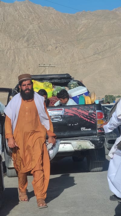 The image shows a man standing next to a black pickup truck, which appears to be overloaded with various items, including luggage, containers, and produce. A child is visible in the back of the truck. The man is wearing an orange shalwar kameez, a brown cap, and sandals. He has a long beard and is holding something in his hand. The background shows a mountainous landscape under a clear blue sky. The truck's license plate is partially visible. The rear of the truck has a red and black tiger stripe design with the name "Hilux Vigo" written on it. The location is identified as Bostan, Pakistan.