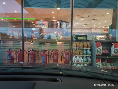 The image captures a view through a large glass window of what appears to be a service station or convenience store. In the foreground, the dashboard of a vehicle is visible, with the windshield reflecting the interior and exterior of the establishment. Reflected in the glass are shelves stocked with various products, including bottles of engine oil and bags of charcoal. There are also fire extinguishers displayed prominently in front of the shelves. Inside the store, a person wearing glasses is visible behind a counter, appearing to be an employee. A menu board and promotional displays are mounted on the wall. The overall impression is one of a busy retail environment within a service station setting. The timestamp "13.06.2026 05:39" indicates the date and time the photograph was taken. The location is identified as Kalugerovo, Bulgaria.