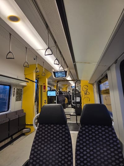 The image displays the interior of a public transit train, located in Neulußheim, Germany. The perspective is from behind two empty, dark grey fabric seats with a repeating polka-dot pattern, facing forward down the central aisle.
The cabin features a primary color scheme of bright yellow and white/light grey. Overhead, multiple white rectangular fluorescent light panels illuminate the space, complemented by a single circular, warm-toned light source on the upper left ceiling. Numerous silver-colored hanging grab handles are suspended from the ceiling. Several digital display screens are mounted throughout the cabin; one screen in the mid-left displays white text on a dark background.
Along the left side of the cabin, a row of five additional patterned seats is visible, adjacent to large rectangular windows that show a blurred, dark exterior. A yellow-framed door or partition is also present on the left. On a yellow panel along the right side of the cabin, yellow spray-paint graffiti, including letters resembling "SC" or "GSC", is visible. The floor throughout the cabin is light grey. Further down the aisle, the cabin structure indicates a possible transition to a lower level or articulated section, with additional seating and partitions extending into the background. No individuals are present in the depicted scene.