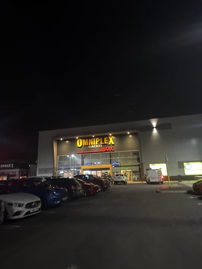 The image displays an exterior nighttime scene of a commercial complex situated in Craigavon, United Kingdom. The dominant structure is the Omniplex Cinemas Maxx building, a modern, multi-story facility with a grey facade and a wide glass entrance. The building's illuminated sign prominently features "OMNIPLEX" and "MAXX" in yellow, with "CINEMAS" in smaller red text. Multiple wall-mounted uplights and downlights illuminate the facade, and interior lights are visible through the glass entrance.
In the foreground and mid-ground, a dark asphalt parking lot contains numerous parked passenger vehicles. These include a white Mercedes-Benz (partially visible front, license plate '09D8 LJW'), a blue car, several dark-colored vehicles, and a red car. A white SUV is parked closer to the cinema entrance, facing away. To the right, a white commercial van equipped with roof racks is parked, near a yellow bollard and other illuminated signage fixtures. To the immediate left of the Omniplex building, a section of another commercial establishment, identified by partial signage as "CONNER'S," is visible with its interior lights on. The sky above is dark, consistent with nighttime conditions. No individuals or specific actions are discernible within the frame.