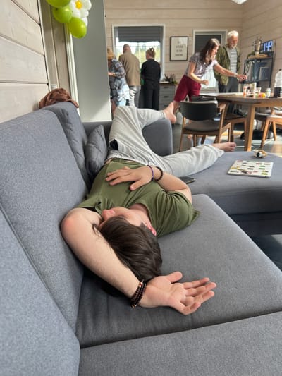 A young male is asleep on a grey couch with his head resting on his arm. He is wearing a green t-shirt and light grey sweatpants. He has brown hair and is wearing bracelets on both wrists. In the background, reflected in a mirror, are several adults standing in a kitchen and dining area. There is a table with chairs, and a woman in a floral shirt and red pants appears to be dancing. The location is Svetloe, Russia.