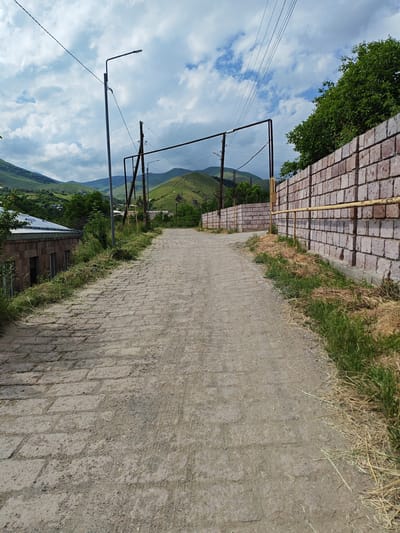 A vertical, low-angle shot captures a cobbled path ascending through a rural Armenian village. The path, paved with light-colored, rough-hewn stones, dominates the foreground and midground, gradually narrowing as it curves slightly to the right and disappears into the distance. Sparse patches of dry grass and weeds sprout along the edges of the path, contrasting with the rough texture of the stones.

To the left of the path, a building with stone walls and a metallic roof is visible, partially obscured by overgrown vegetation. Further up the path, utility poles with electrical wires crisscross the sky, accompanied by a street lamp. A low wall constructed of light pink bricks runs along the right side of the path, topped with a yellow pipe. Behind this wall, a dense canopy of green trees suggests a natural landscape beyond.

The background features rolling green hills, characteristic of the Bazum region of Armenia. The sky above is a mix of blue and white, with fluffy clouds that suggest a bright but possibly overcast day. The overall impression is one of a serene, rural setting, likely in the late morning or early afternoon given the lighting. No people or specific activities are depicted. There is no visible text in the image.