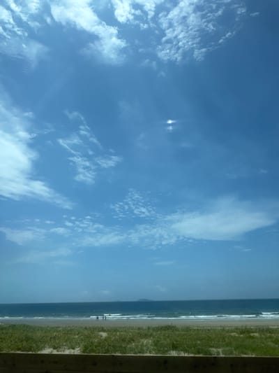 The image depicts a coastal scene, likely Anmyeon-eup, South Korea, under a vast blue sky dotted with white clouds. The foreground features a patch of green grass and sand, bordered by a wooden structure. Beyond this, the ocean stretches out, with gentle waves rolling onto the sandy beach. In the middle ground, several figures are visible near the water's edge, their specific actions indistinct. In the distance, a small island or landmass is faintly visible on the horizon. The overall impression is a serene beach environment on a clear day.