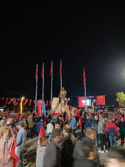 The image shows a large gathering of people at night in Silivri, Türkiye. The location is confirmed by the coordinates provided (41.072, 28.244), which correspond to a public square in the city. The scene is illuminated by streetlights and the people are gathered around a monument. The monument appears to be a statue of a man standing on a raised platform. Several Turkish flags are flying from flagpoles around the monument. Some of the people in the crowd are holding Turkish flags. There are many people standing and interacting with each other in the foreground. The atmosphere appears to be celebratory, possibly a patriotic gathering.