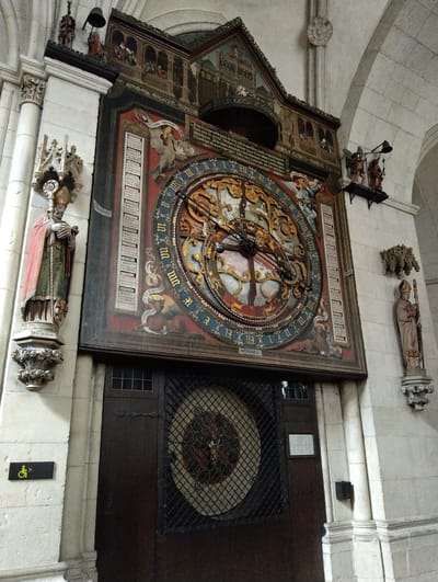 The image captures the astronomical clock of Münster Cathedral, located in Münster, Germany. The clock is prominently displayed above a dark wooden door that is likely an entrance or storage area, a sign for handicapped accessibility is adjacent to it. The clock is highly ornate, with a complex circular design featuring zodiac symbols, religious figures, and inscriptions. Two statues of religious figures flank the clock on either side, mounted on ornate corbels, a bell and bell ringer are located on top of a pillar to the left. The clock is integrated into the cathedral's architecture, with the structure and decorative elements harmonizing with the surrounding stonework. The environment consists of light-colored stone walls with arched openings.