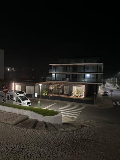 A nighttime urban scene showcases a modern multi-story building with illuminated windows. A crosswalk spans the street, leading to the building. A white van is parked nearby, adjacent to a "Do Not Enter" sign and a streetlamp. The ground is covered with small, grey cobblestones on one side and asphalt on the other. Distant streetlights line the road, disappearing into the dark horizon. A small patch of green grass is nestled between the sidewalk and the street.
