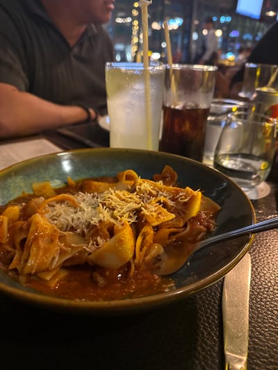 The image displays a dining scene at night, featuring a close-up of a meal on a dark, textured table. A dark teal ceramic bowl in the foreground contains wide, flat pasta (likely pappardelle) coated in a reddish-brown sauce and topped with grated cheese. A silver fork is inserted into the pasta. To the right of the bowl, a silver dining knife rests on the table. Behind the pasta bowl, from left to right, are a tall clear glass holding a light, opaque, yellowish-green beverage with ice and a white straw; a clear glass containing a dark brown carbonated beverage; and a stemmed glass with clear water. In the upper-left, a person wearing a dark, short-sleeved collared shirt is partially visible, facing right. The background is out of focus, showing numerous blurred circular light sources, indicative of decorative lighting in a dimly lit evening restaurant or bar setting. No specific landmarks or signage are present to identify the precise location within San Agustín, Mexico.