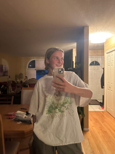 A young, fair-skinned person with short blonde hair is holding a smartphone to take a selfie in what appears to be the interior of a home. The person is wearing a white t-shirt with a floral design and what looks like grey pants. The background suggests an interior hallway or entrance area with a front door visible. There's a chandelier-type light fixture above, and a wooden floor can be seen leading away from the entrance. In the reflection, a dining area with furniture and decorative items can be observed. Determining the specific location within Kelowna, Canada from the image alone is not possible without additional information or context.