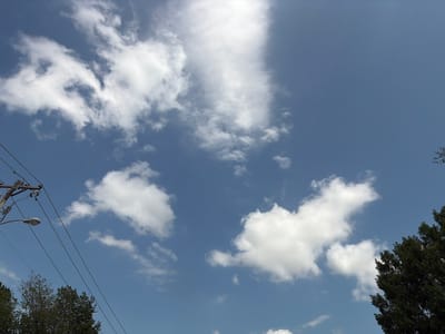 The image depicts a clear blue sky with scattered white clouds. In the lower left corner, a utility pole with power lines and a street lamp is visible, extending diagonally upwards. The tops of trees can be seen along the bottom edge of the image. The scene is outdoors and appears to be taken during the daytime, possibly in Grapevine, Texas, United States of America. There are no people or animals present in the frame.