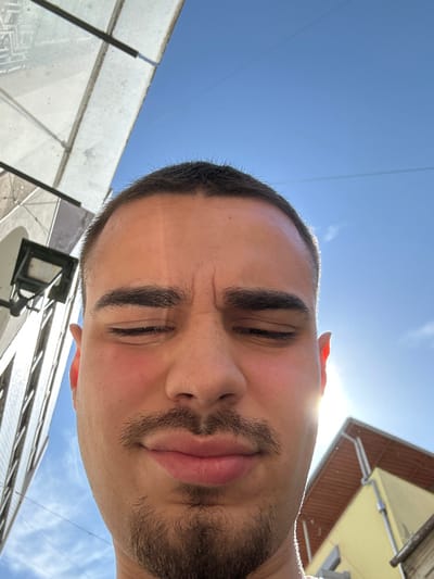 A low-angle close-up photograph captures a male individual with short dark hair, dark eyebrows, a mustache, and a goatee. His eyes are squinted shut, and his forehead is furrowed, suggesting a reaction to bright light. His cheeks exhibit a reddish flush. Strong direct sunlight, visible as a glare, originates from the lower right of the frame, illuminating the subject's right side. The background features a clear blue sky. To the left, a multi-story building with reflective surfaces or windows and an attached street lamp under a glass canopy structure is partially visible. To the right, a yellow-facade building with a brown-tiled roof and a gutter system is observed. Faint utility lines traverse the upper sky. The scene is situated in the city of Chaves, Portugal.