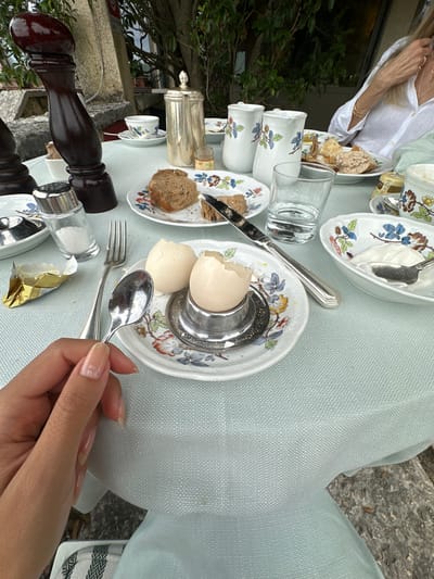 An overhead shot captures a table set for breakfast outdoors, possibly on a terrace overlooking greenery and stonework. In the foreground, a woman's hand with light-colored, long nails holds a spoon, poised over a plate with a cracked-open hard-boiled egg in a silver egg cup. The plate is adorned with a floral pattern. Next to it, a fork rests on the light blue, textured tablecloth. Other items on the table include a salt shaker, a piece of butter in gold foil, a small jar of jam, a glass of water, a coffee pot, a teacup, two pitchers, and plates of bread and what appears to be yogurt or cream. A person in a white shirt is visible in the background. The overall scene suggests a leisurely breakfast in a serene setting, possibly within a villa or garden in Maser, Italy, given the aesthetic and lush surroundings.