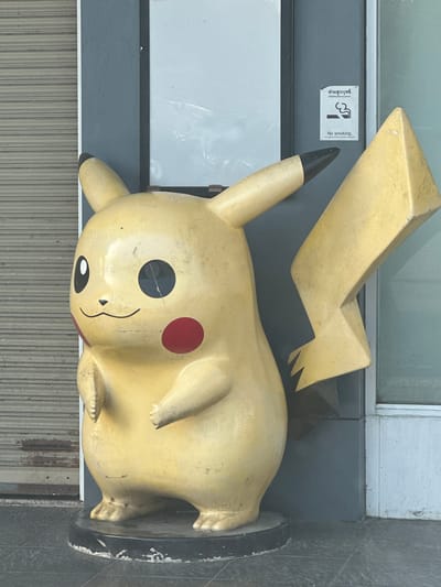 A weathered, life-size yellow statue of Pikachu stands on a black circular base on a tiled walkway in front of a building with a gray facade and a roll-up shutter door. The statue has visible signs of age and wear, with scratches and dirt marring its surface. To the right of the statue, on the gray wall, a "No Smoking" sign is posted, featuring a graphic of a cigarette with a red line through it and text in both Thai and English. The background shows the muted tones of urban architecture, with the shutter door on the left and parts of the building's exterior on the right, including a glass pane. The lighting suggests it is daytime, with shadows cast by the statue. The location is identified as Ban Ko Pru, Thailand.