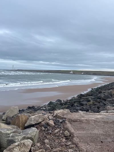 The image displays a coastal scene at Montrose, United Kingdom, specifically looking across Montrose Bay. The sky is entirely overcast with dull grey clouds. In the foreground, large, irregular dark grey and brown rocks, consistent with riprap, dominate the lower left and center, bordering a dark, rough ground surface that occupies the lower right. The midground features choppy, dark grey-green seawater with white breaking waves rolling onto a wide, crescent-shaped sandy beach, which appears dark and wet near the water's edge and lighter further inland. In the distance on the far left, a tall, slender white lighthouse is visible on a low headland. To the right, across the bay, another, shorter white lighthouse with a yellow cap, accompanied by smaller white buildings, stands on a grassy headland, from which a low sea barrier extends into the water. No individuals, vehicles, or specific wildlife are discernible within the scene.