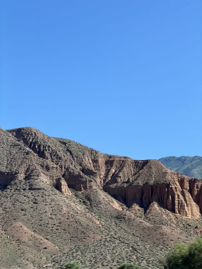 This is a landscape photograph of a mountainous region under a clear blue sky. The foreground features a rugged, reddish-brown mountain slope sparsely covered with dry vegetation. The mountain displays intricate geological formations, including vertical striations and eroded cliff faces, suggesting a history of weathering and erosion. In the background, further mountain ranges with a greener hue are visible, indicating a larger, natural environment. The image appears to be taken during daylight, with the sun casting shadows that highlight the textures of the terrain. The scene is devoid of human presence or any man-made structures, emphasizing the wild and natural character of the location. This geographical area is identified as being in the vicinity of Kok Moynok Eki, Kyrgyzstan.
