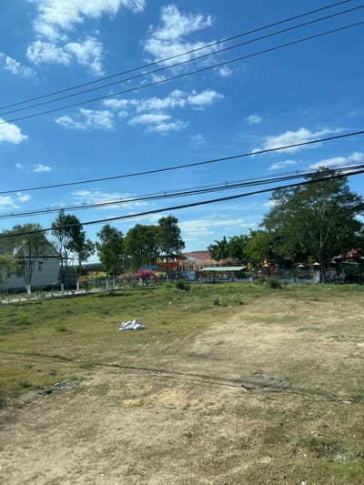 A blue sky with scattered clouds is traversed by electrical wires. A grassy, partially dried field occupies the right side of the image. A building peeks out from behind trees and a fence, with some colorful decorative elements visible behind the fence. A white object lies on the field.