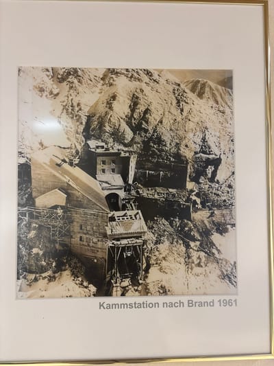 The image is a framed black and white photograph of a mountainous landscape with a large building complex under construction. The building appears to be a station, possibly for a cable car or other transport system, situated on a steep, rocky mountainside. There are scaffolding and construction materials visible, suggesting an ongoing building project. The architecture of the station is robust and functional, with multiple levels and windows. The surrounding environment is rugged and arid, with prominent rock formations and sparse vegetation. The photograph is captioned "Kammstation nach Brand 1961," which translates to "Summit station after fire 1961." This suggests the image depicts the reconstruction of a summit station in Brand, Austria, following a fire in 1961. While the prompt mentions Ehrwald, Austria, the caption specifically refers to Brand.