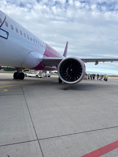 An airport tarmac at Künersberg, Germany, features a close-up view of the engine and fuselage of a pink and white passenger airplane. The airplane's landing gear is visible, as is a ground support vehicle and a boarding ramp. A group of passengers, some carrying luggage, stand near the aircraft, seemingly waiting to board. The sky above is filled with white, scattered clouds. The tarmac is concrete with yellow and red painted markings.