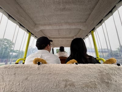 The image shows the interior of a covered vehicle, likely a small tour bus or similar transport, with three people visible. The perspective is from the rear of the vehicle, looking forward. The ceiling and some interior panels are covered in a fuzzy, light-colored material. The sides of the vehicle have transparent mesh panels with a grid pattern, allowing views of the exterior, which appears to be a park or natural setting with trees. Bright yellow support poles are visible within the vehicle.

In the foreground, a fluffy, cream-colored fabric covers the back of a seat. Two small, fuzzy, brown stuffed animal-like objects are attached to the top of this seat, one on the left and one on the right.

Immediately in front of this seat, facing forward, are two passengers. On the left is a person wearing a white collared shirt and a black baseball cap. They are wearing glasses and appear to be looking forward. On the right is a person with long, dark hair, wearing a black top. They are also facing forward.

Further ahead, in the middle of the vehicle, is a third person, also facing forward. This person is wearing a hooded jacket and a baseball cap, with their back to the camera.

The scene is set in the city of Nagakute, Japan, likely within the Toyota Shirakawa Nature Park or a similar recreational area, given the foliage and the type of transport commonly found in such parks. The overall impression is one of leisure travel in an outdoor, park-like environment.