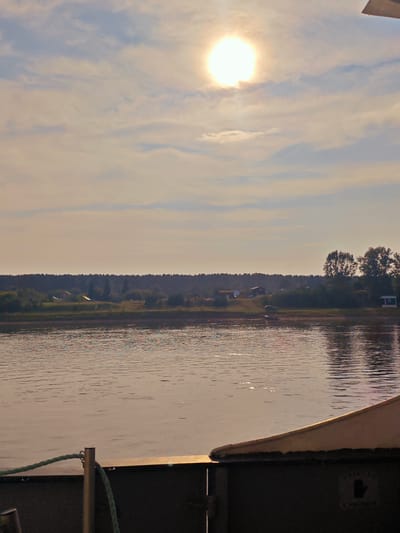The image depicts a wide body of water reflecting the sky and the sun. The sun, a bright orb, is visible in the upper portion of the sky, partially obscured by soft clouds. On the far bank of the water, a line of trees forms a dense, dark border. In front of the trees, there are scattered small buildings and structures, suggesting a rural settlement. A few taller trees stand out amongst the foliage. In the foreground, the edge of a vessel is visible, with a metal railing and a green rope. The water's surface is rippled, with highlights from the sunlight. The scene appears to be set in the village of Yukseevo, Russia, given the presence of a natural landscape and likely a river or lake. There are no people or overt actions depicted. The environment is tranquil and natural, with a hint of human habitation on the horizon.