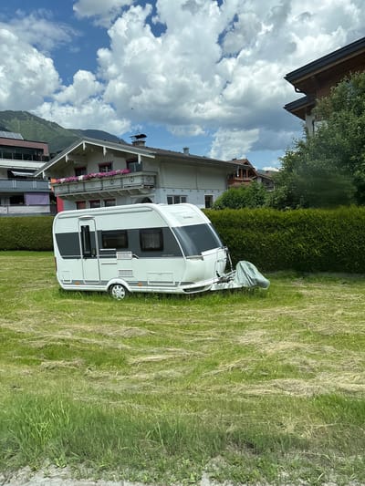 This image, taken in Bruck an der Großglocknerstraße, Austria, depicts a white and gray caravan parked on a grassy lawn. The caravan has tinted windows and a brand name "Hobby" visible on its side. A light green cover is partially draped over the front of the caravan. Behind the caravan, a tall, neatly trimmed green hedge separates it from a white building with dark trim and balconies adorned with pink flowers. The building appears to be a house or a guesthouse, with visible architectural details like a wooden roof structure and decorative elements on the balconies. In the background, a lush green mountain slope rises under a partly cloudy blue sky, suggesting a daytime scene with fair weather. The overall setting is serene and picturesque, typical of a mountain resort town. No people are visible in the image.